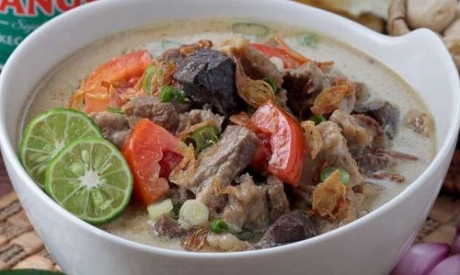 Satu mangkuk putih berisi soto Betawi bersantan yang dilengkapi daging sapi, tomat, daun bawang, bawang goreng, dan jeruk nipis. 